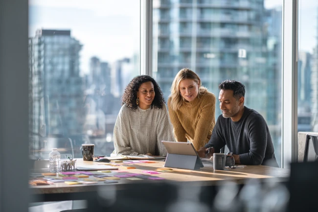 Three professionals collaborating over a laptop in a high-rise office, representing Remote Ops Co.’s elite remote support and operations team.